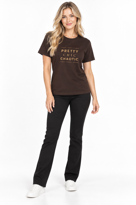 A woman modeling a dark brown short-sleeve graphic t-shirt with gold text that reads 'PRETTY CHILLY CHAOTIC' on the front, paired with black pants and white sneakers, against a plain white studio background.