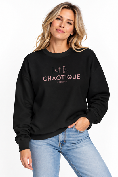 A woman wearing a black crewneck sweatshirt with the words 'L'art Du CHAOTIQUE' printed in pink on the front, paired with light blue jeans, against a plain white background.