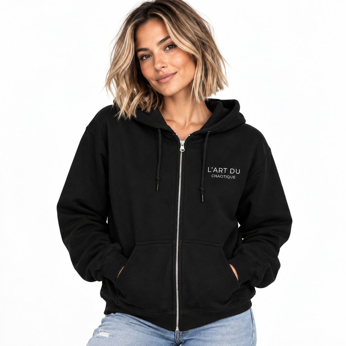 A woman wearing a black zip-up hoodie with 'L'ART DU CAOTIQUE' printed in white on the left chest, paired with light-wash blue jeans, set against a plain white background.