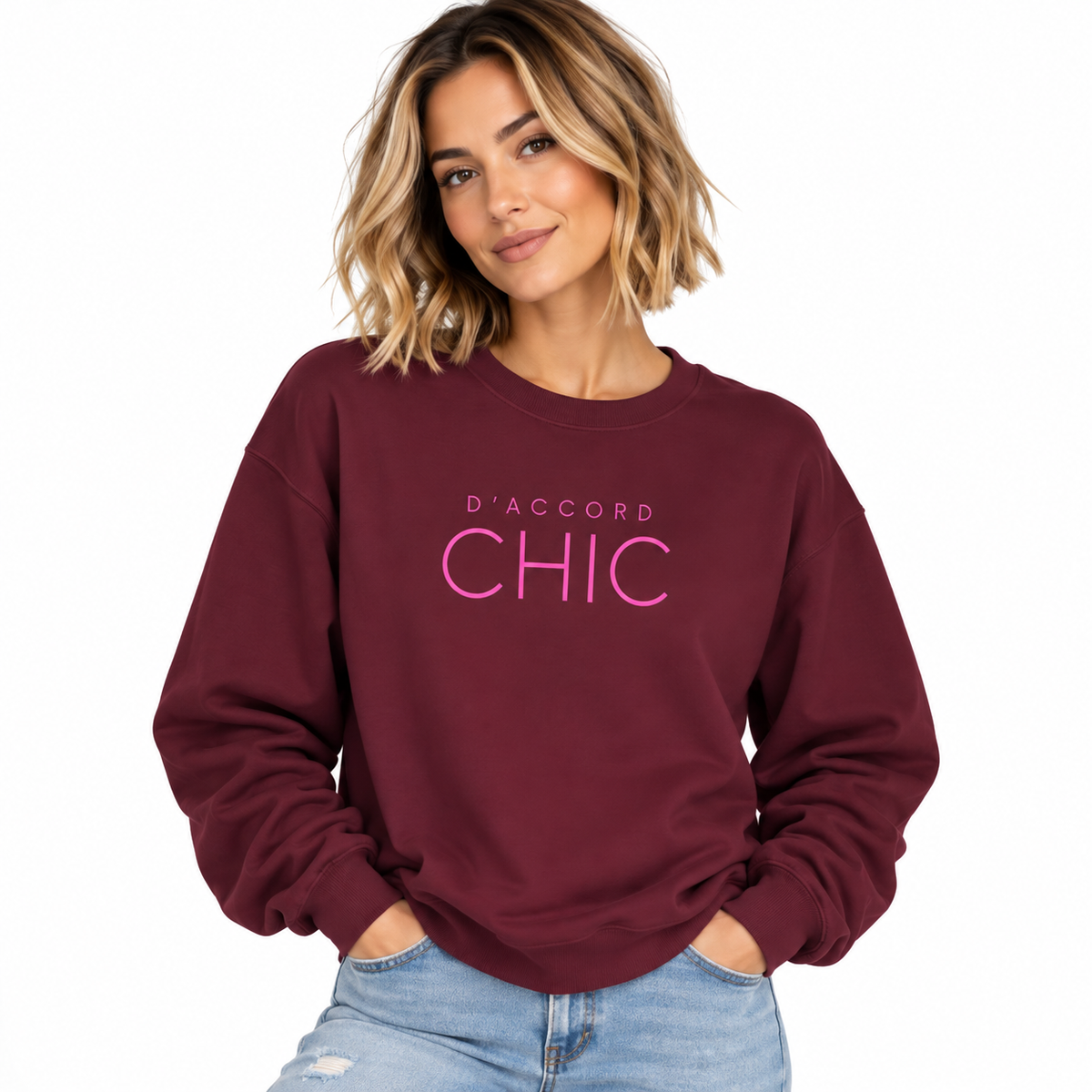 A woman wearing a maroon crewneck sweatshirt with 'D'ACCORD CHIC' printed in pink across the chest, paired with light blue jeans, against a plain white background.