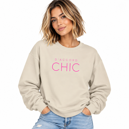 A woman wearing a light beige crewneck sweatshirt with 'D'ACCORD CHIC' printed in pink across the chest, paired with light blue jeans, set against a plain white background.
