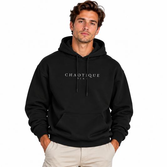 A man wearing a black pullover hoodie with 'CHAOTIQUE BAEU' printed in white across the chest, paired with light beige pants, set against a plain white background.