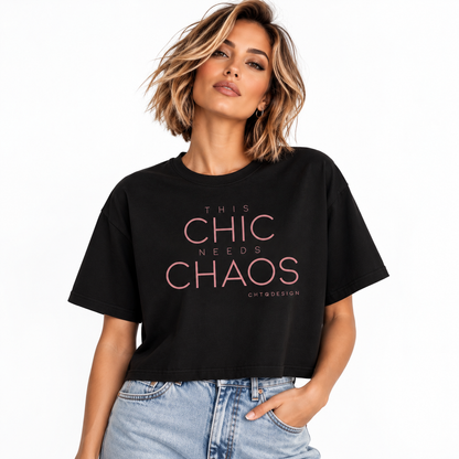 A woman wearing a black cropped graphic t-shirt with the text 'THIS CHIC NEEDS CHAOS' in pink, paired with light-wash blue jeans, against a plain white background.