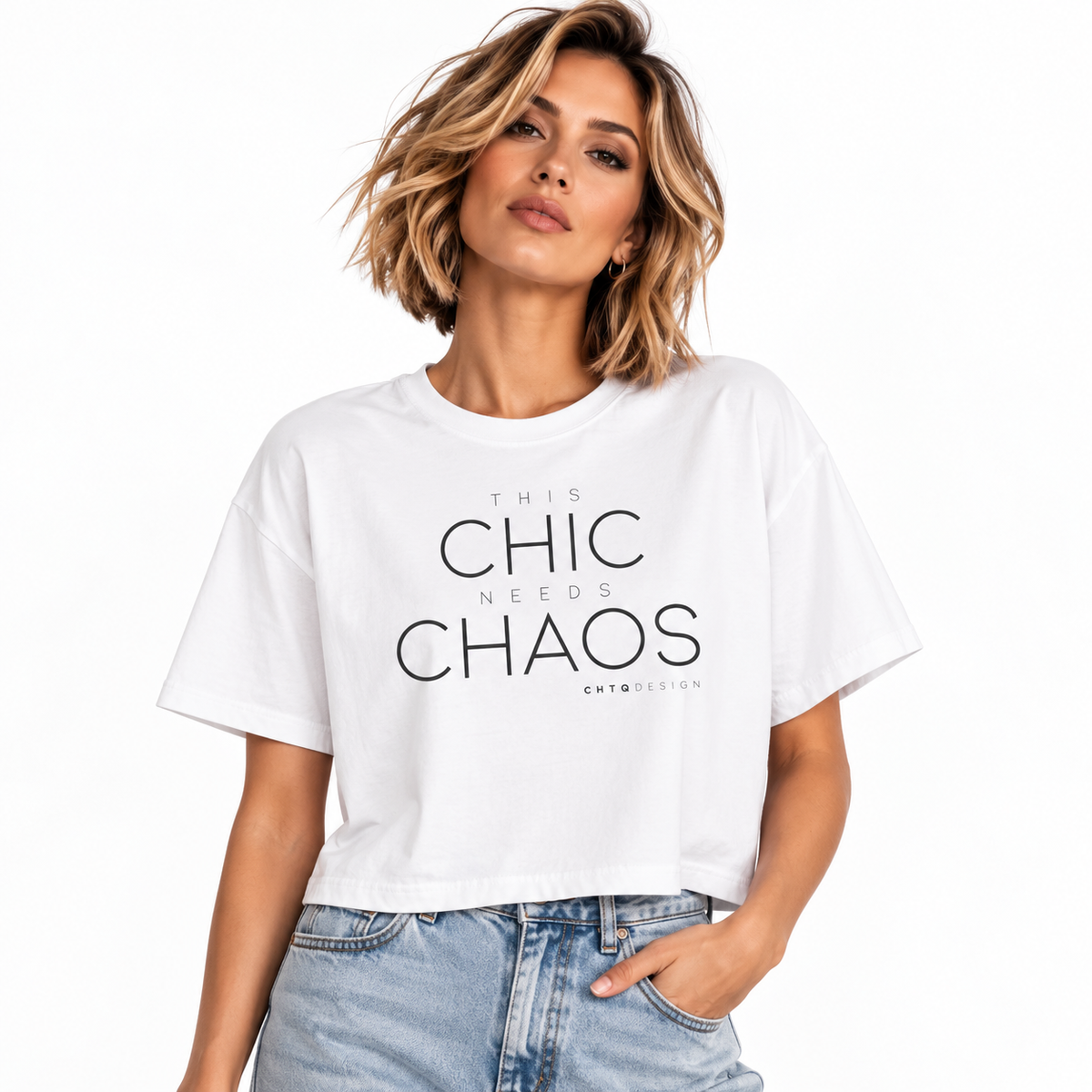 A woman wearing a white cropped graphic t-shirt with the text 'THIS CHIC NEEDS CHAOS' in black, paired with light-wash blue jeans, against a plain white background.