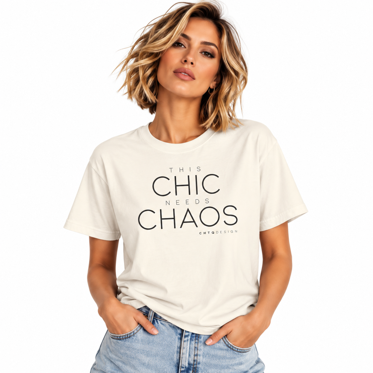 A woman with short blonde hair wearing a cream-colored graphic t-shirt with the text 'THIS CHIC NEEDS CHAOS' in black, paired with light-wash blue jeans, against a plain white background.