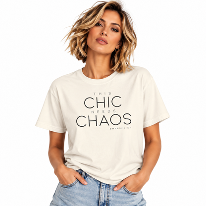 A woman with short blonde hair wearing a cream-colored graphic t-shirt with the text 'THIS CHIC NEEDS CHAOS' in black, paired with light-wash blue jeans, against a plain white background.