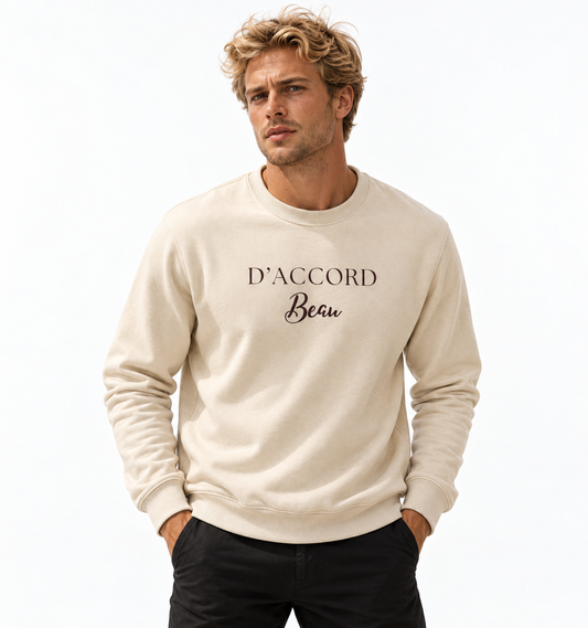 A man wearing a cream-colored crewneck sweatshirt with 'D'ACCORD Beau' printed in black across the chest, paired with black pants, against a plain white background.