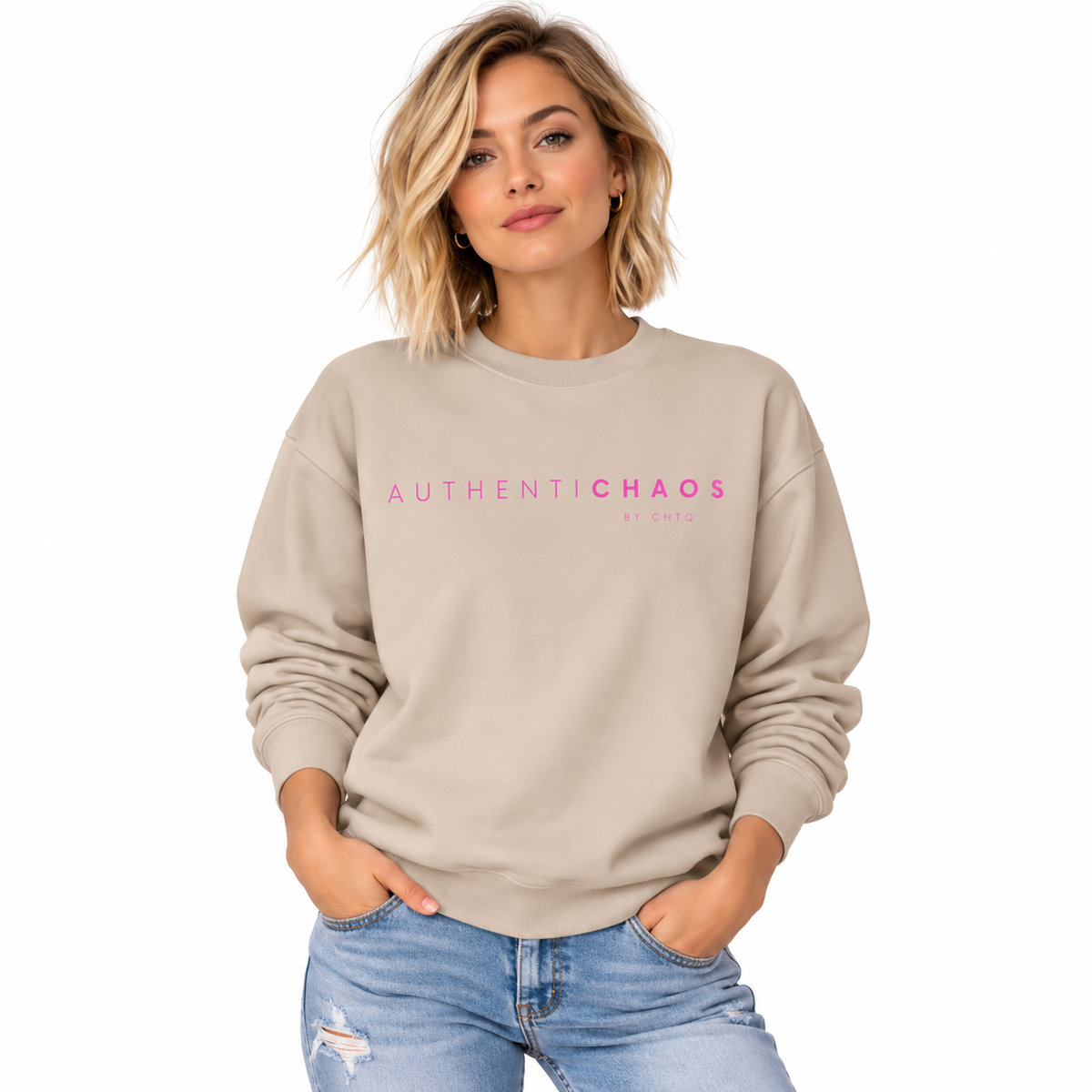 A woman wearing a beige crewneck sweatshirt with 'AUTHENTICCHAOS' printed in pink across the chest, paired with light blue jeans, set against a plain white background.