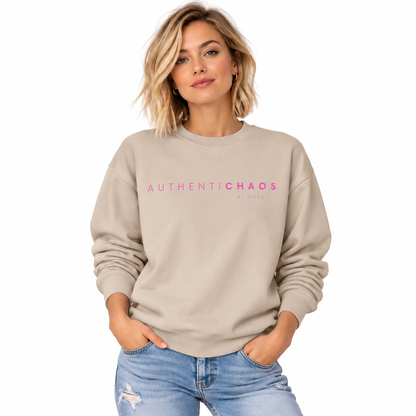 A woman wearing a beige crewneck sweatshirt with 'AUTHENTICCHAOS' printed in pink across the chest, paired with light blue jeans, set against a plain white background.