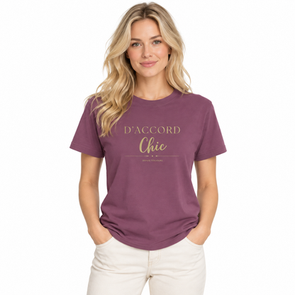 A woman with blonde hair wearing a mauve-purple t-shirt with gold text that reads 'D'ACCORD Chic' and white pants, standing against a plain white background.