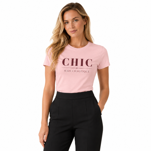 A woman modeling a light pink crewneck t-shirt with 'CHIC MAIS CHAOTIQUE' printed in dark pink on the front, paired with black trousers, against a plain white background.