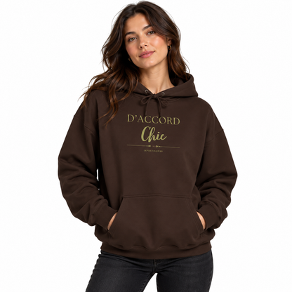 A woman wearing a dark brown pullover hoodie with 'D'ACCORD Chic' printed in gold on the chest, paired with black jeans, against a plain white background.