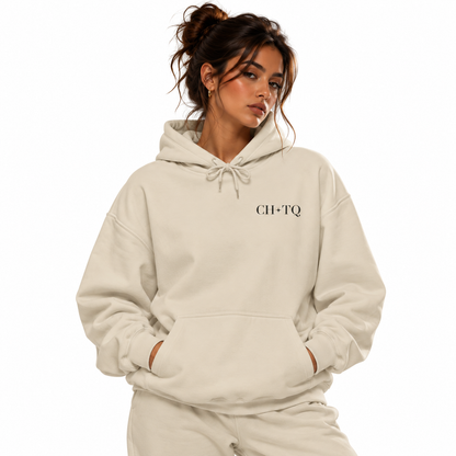 A woman wearing a light cream-colored hoodie with 'CH-TQ' printed in black serif font on the left chest, paired with matching sweatpants, set against a solid white background.