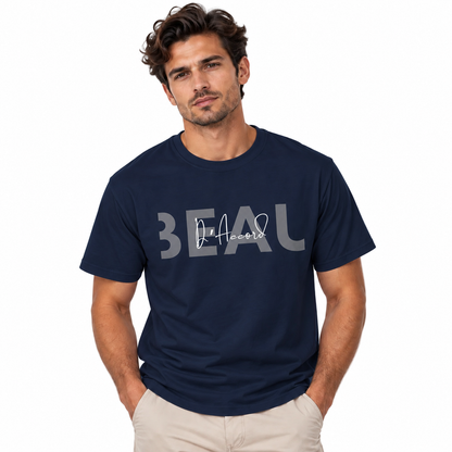A man wearing a Navy blue BEAU D’Accord graphic T-shirt featuring large grey BEAU lettering with white handwritten D’Accord script across the chest, paired with beige pants, against a plain white background.