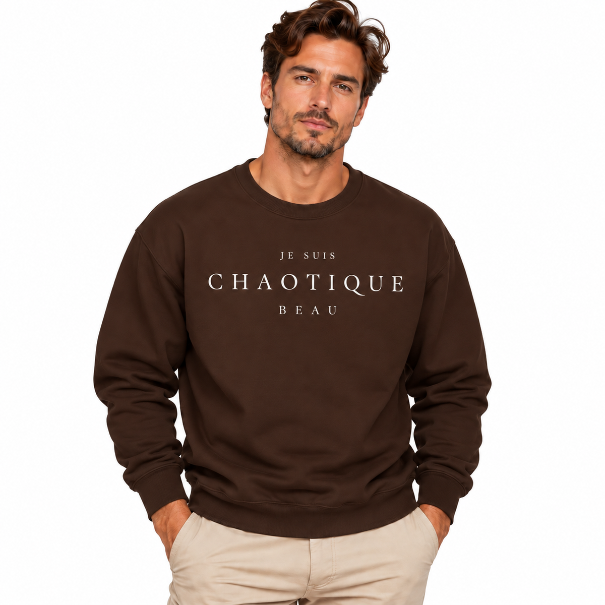 A man wearing a brown crewneck sweatshirt with the text 'JE SUIS CHAOTIQUE BEAU' printed in white across the chest, paired with beige pants, set against a plain white background.