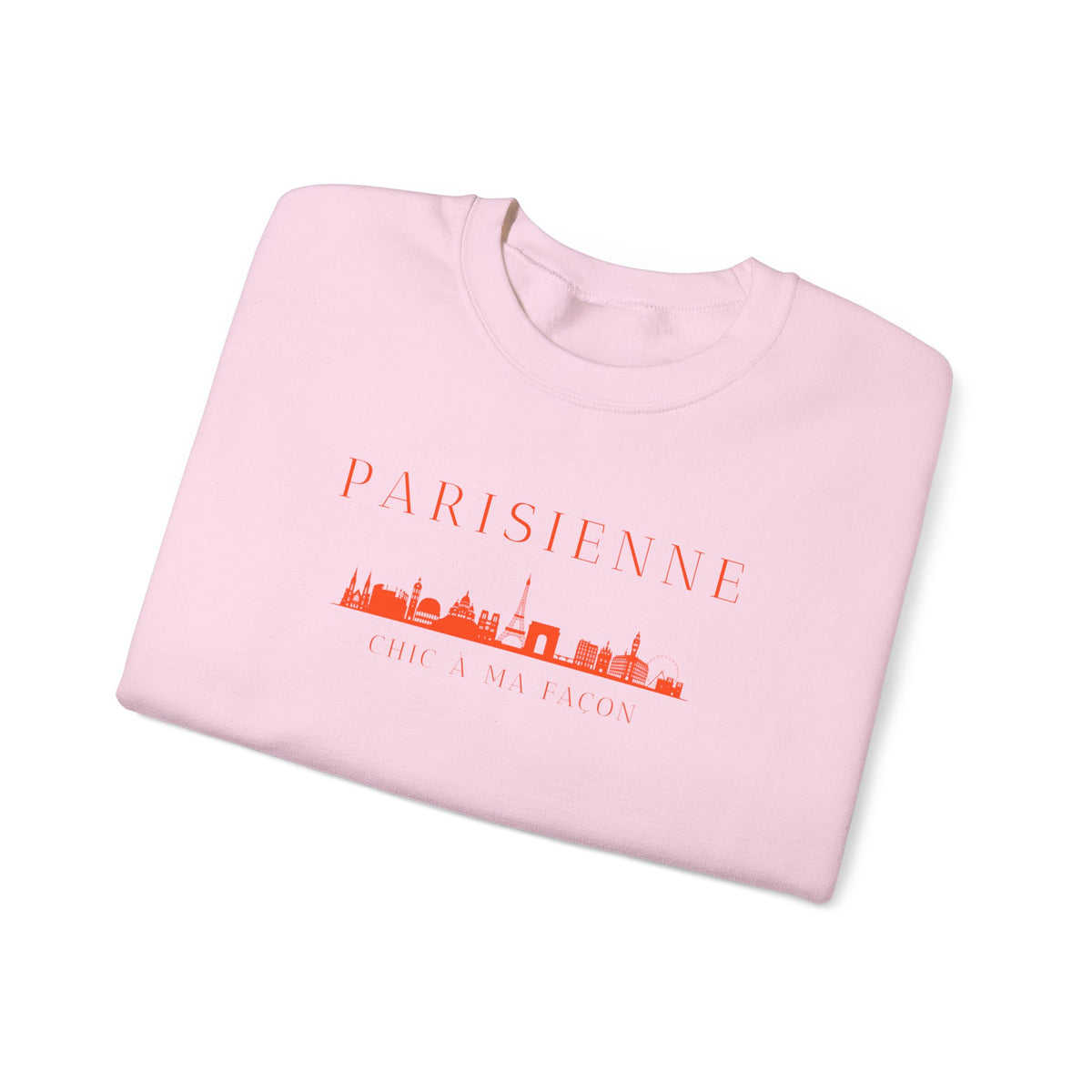 A folded light pink crewneck sweatshirt with a bright orange “PARISIENNE” graphic, Paris skyline design, and “CHIC À MA FAÇON” text on the chest, displayed against a clean white background.
