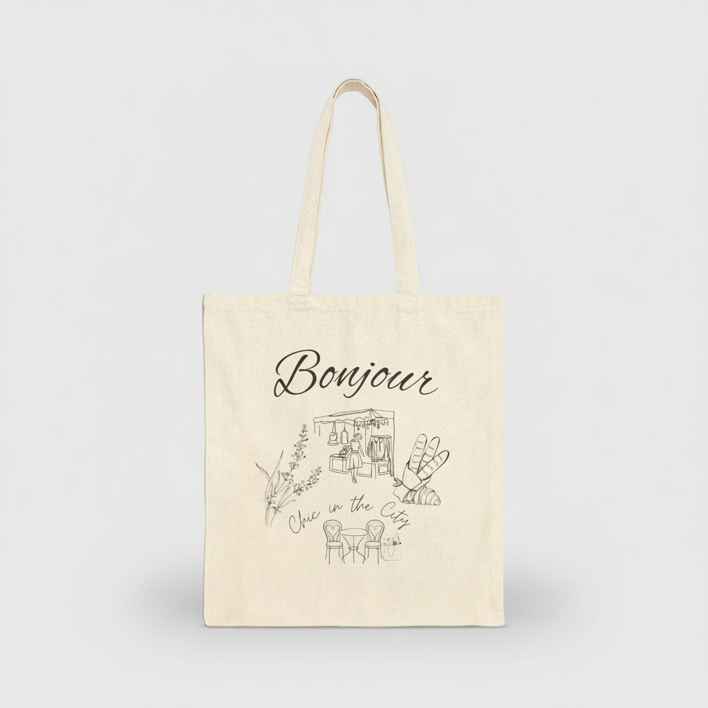 An off-white canvas tote bag with long handles, featuring a black line art illustration of a cafe and the word 'Bonjour' printed on the front, set against a plain white background.