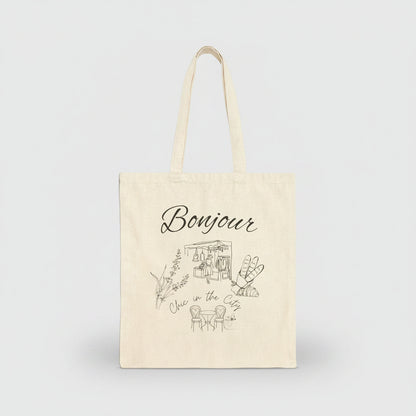 An off-white canvas tote bag with long handles, featuring a black line art illustration of a cafe and the word 'Bonjour' printed on the front, set against a plain white background.