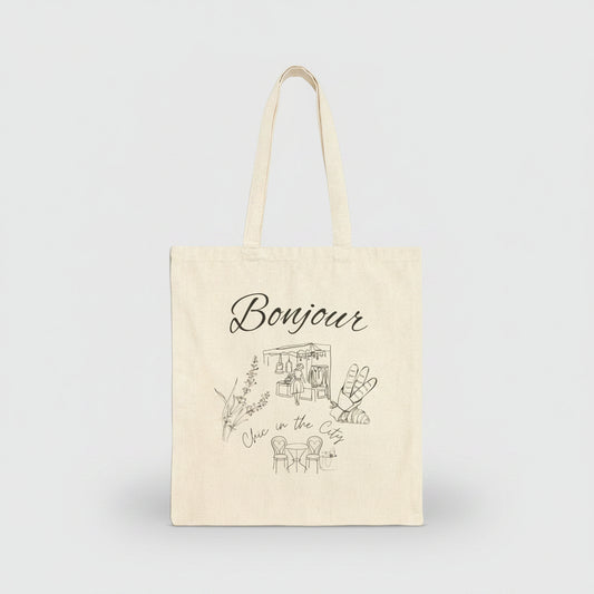 An off-white canvas tote bag with long handles, featuring a black line art illustration of a cafe and the word 'Bonjour' printed on the front, set against a plain white background.