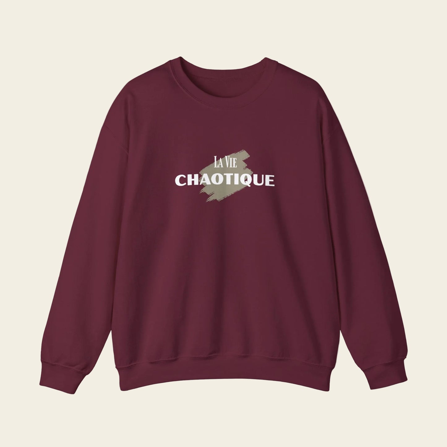 A maroon crewneck sweatshirt with a graphic on the chest that reads 'LA VIE CHAOTIQUE' in white text over a muted olive green paint-splatter shape, displayed on a clean white background.