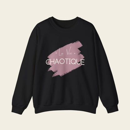 A black crewneck sweatshirt featuring a dusty pink paint brush stroke graphic on the chest with the text 'La Vie CHAOTIQUE' in white, displayed against a solid white background.