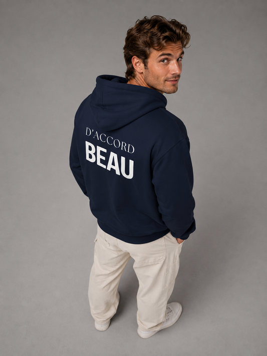 A man wearing a navy blue D’Accord Beau hoodie with bold white back typography, photographed in a light grey studio from a slightly elevated angle while looking back at the camera with a subtle smile.