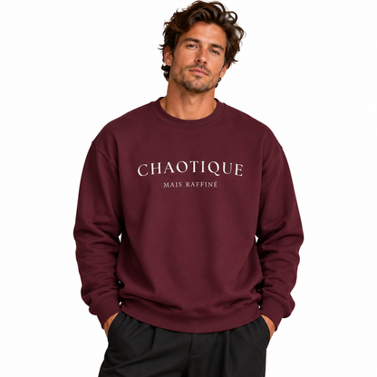 A man wearing a burgundy crewneck sweatshirt with 'CHAOTIQUE MAIS RAFFINÉ' printed in white across the chest, paired with black pants, set against a plain white background.