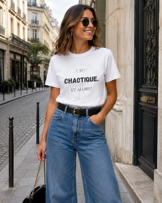 A woman wearing a white graphic T-shirt with black text reading 'CHAOTIQUE. ET ALORS?' and blue wide-leg jeans, standing on a cobblestone street in a city setting.