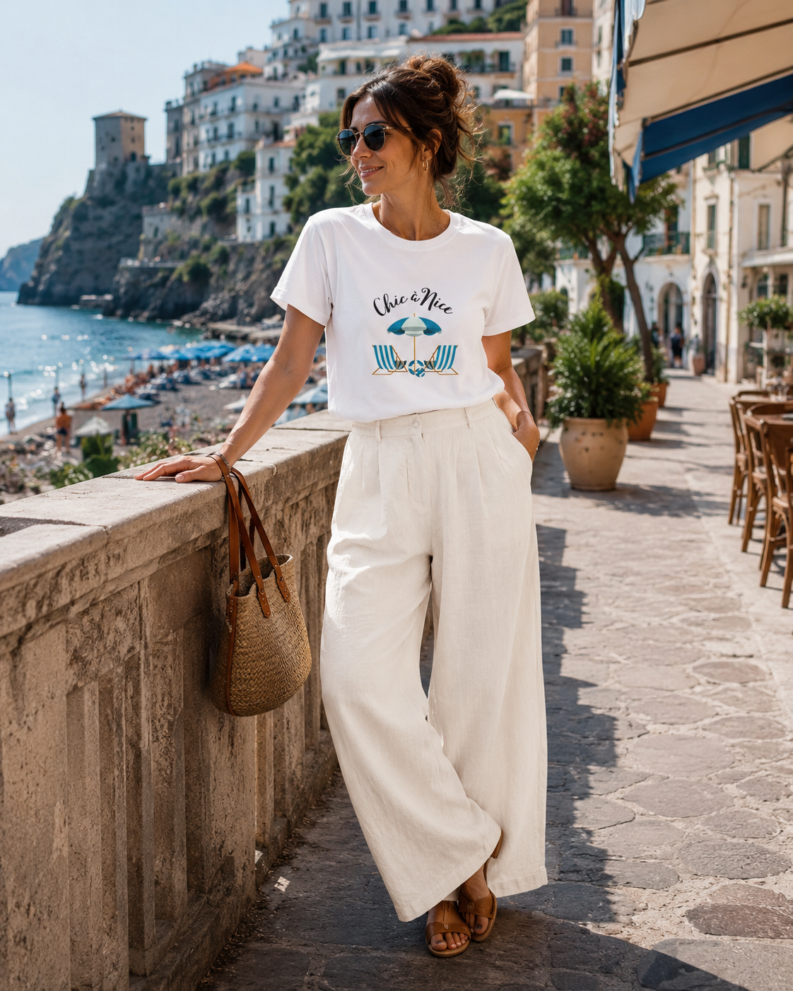 A woman stands on a seaside promenade wearing a white graphic T-shirt and white wide-leg pants, with a woven shoulder bag and sunglasses. Buildings, beach, and sea are visible in the background.