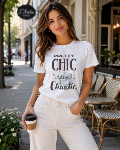 A woman stands on a city sidewalk wearing a white graphic T-shirt with the phrase 'PRETTY CHIC festively Chaotic.' in black lettering, paired with off-white jeans and holding a takeaway coffee cup.