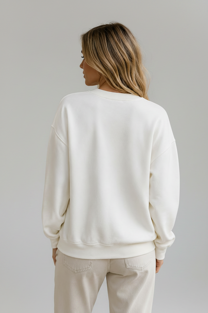 A woman shown from the back wearing a cream-colored crewneck sweatshirt and matching sweatpants, standing in front of a light beige stone wall.
