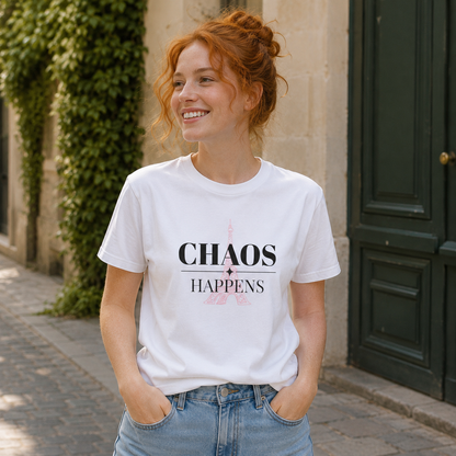 A smiling woman with red hair tied up stands outdoors wearing a white graphic T-shirt with the words 'CHAOS HAPPENS' printed on the front, paired with light blue jeans. The background shows a stone wall with green ivy and a dark green door.