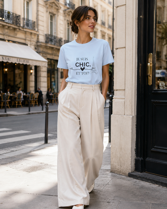 A woman stands on a city sidewalk wearing a light blue graphic T-shirt with the French text 'JE SUIS CHIC. ET TOI?' and cream wide-leg trousers.