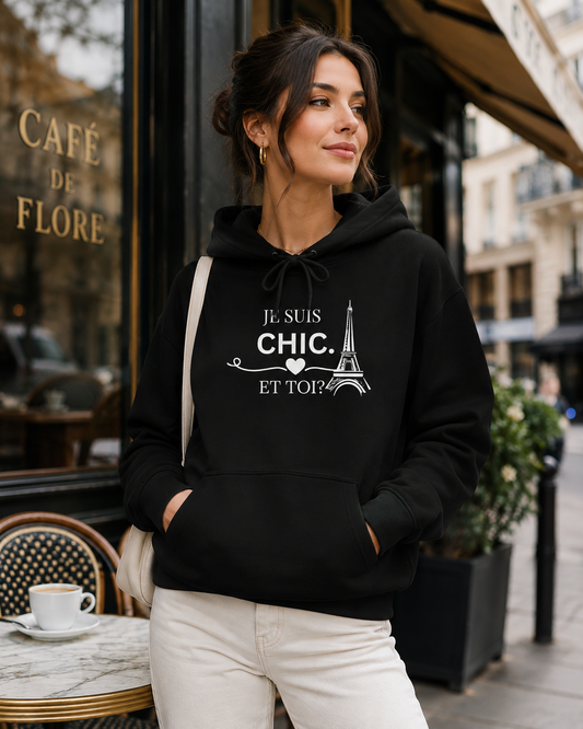 A woman wearing a black hoodie with a white graphic print that reads 'JE SUIS CHIC. ET TOI?' next to a Paris Eiffel Tower illustration, standing on a city street in front of a cafe.