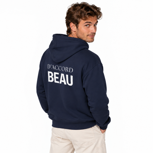 A man wearing a navy blue hoodie with 'D'ACCORD BEAU' printed in white on the back, paired with beige trousers, photographed against a solid white background.