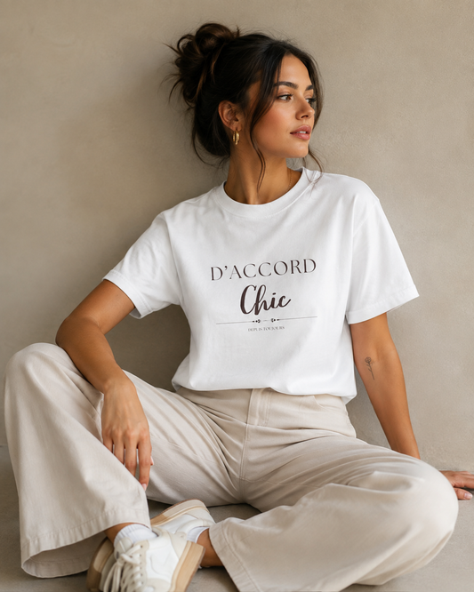 A woman sitting cross-legged against a warm beige studio wall wearing a white D'Accord Chic graphic T-shirt with black typography, paired with beige wide-leg trousers and white sneakers.