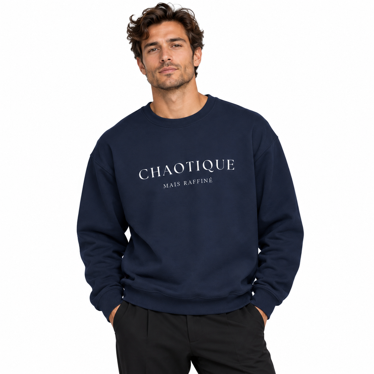A man wearing a navy blue crewneck sweatshirt with 'CHAOTIQUE MAIS RAFFINE' printed in white across the chest, paired with off-white trousers, against a solid gray studio background.