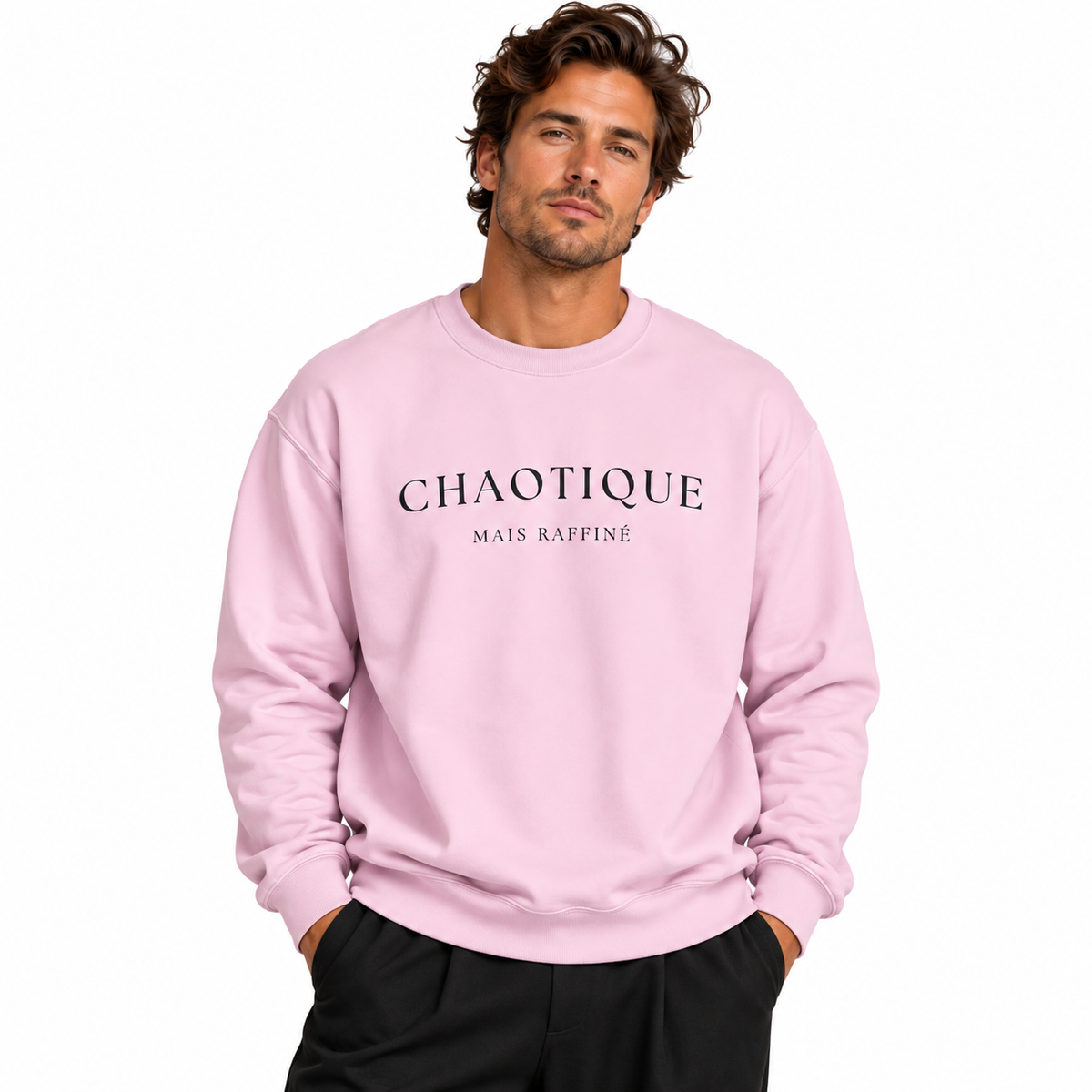 A man wearing a light pink crewneck sweatshirt with 'CHAOTIQUE MAIS RAFFINÉ' printed in black across the chest, paired with black pants, set against a plain white background.