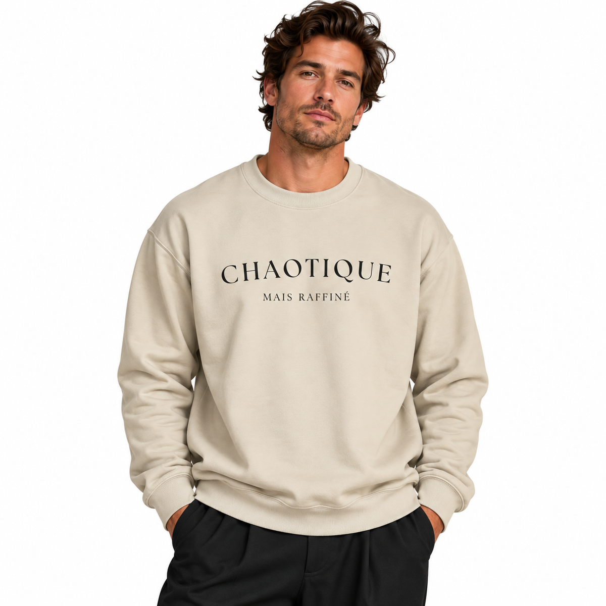 A man wearing a light beige crewneck sweatshirt with 'CHAOTIQUE MAIS RAFFINÉ' printed in black across the chest, paired with black pants, set against a plain white background.