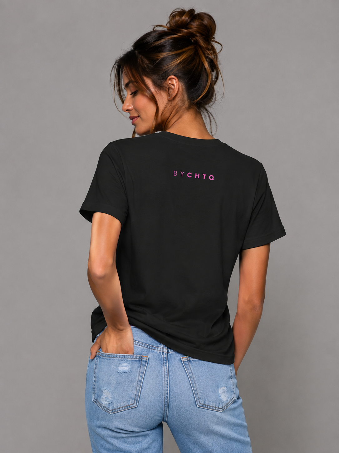 A woman wearing a black short-sleeve T-shirt shown from the back, featuring a small pink 'BY CHTQ' detail below the neckline, paired with light-wash jeans against a neutral gray studio background.