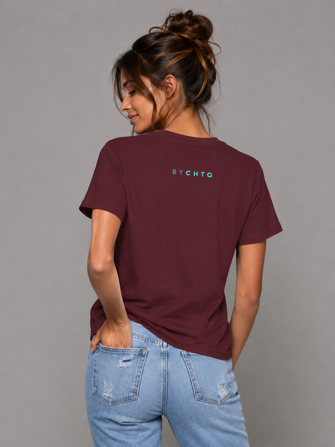 A woman wearing a maroon short-sleeve T-shirt shown from the back, featuring a small turquoise 'BY CHTQ' detail below the neckline, paired with light-wash jeans against a neutral gray studio background.