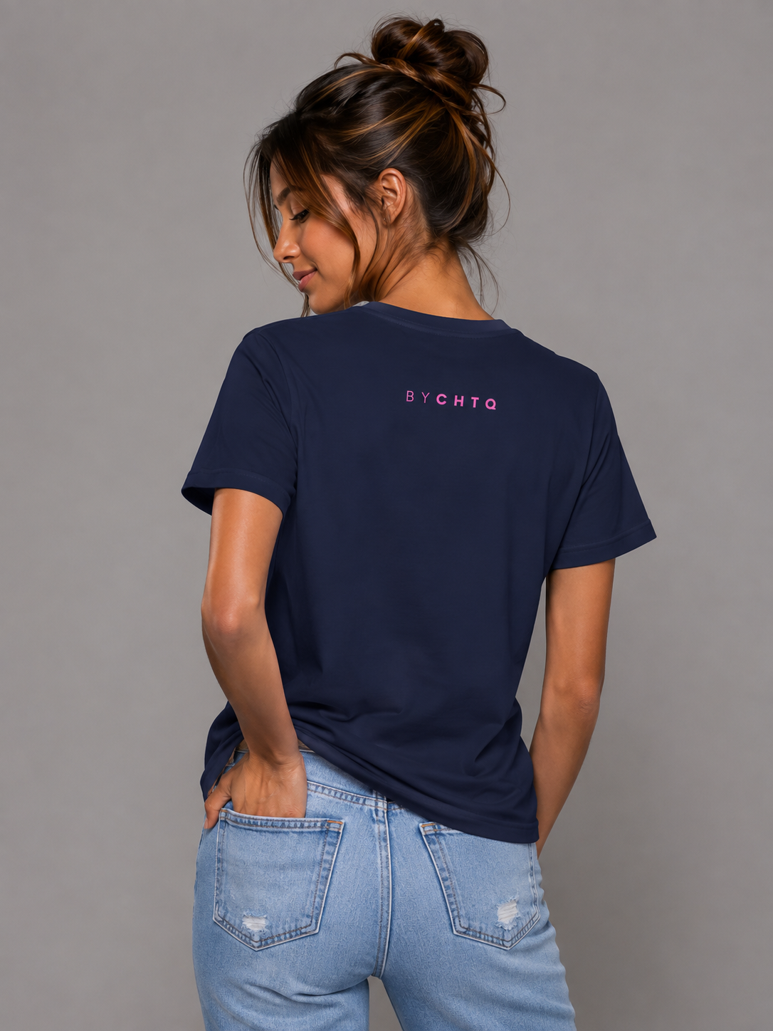 A woman wearing a navy short-sleeve T-shirt shown from the back, featuring a small pink 'BY CHTQ' detail below the neckline, paired with light-wash jeans against a neutral gray studio background.