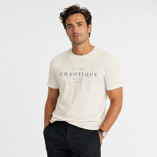 A man wearing a light beige Je Suis Chaotique Beau graphic T-shirt with dark tailored trousers, photographed against a clean white studio background.