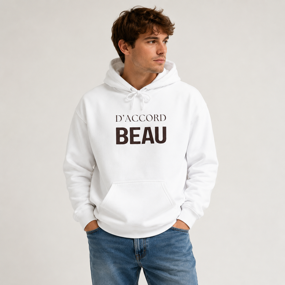 A man wearing a white pullover hoodie featuring black “D’ACCORD BEAU” text across the chest, paired with blue jeans, photographed against a light gray studio background.