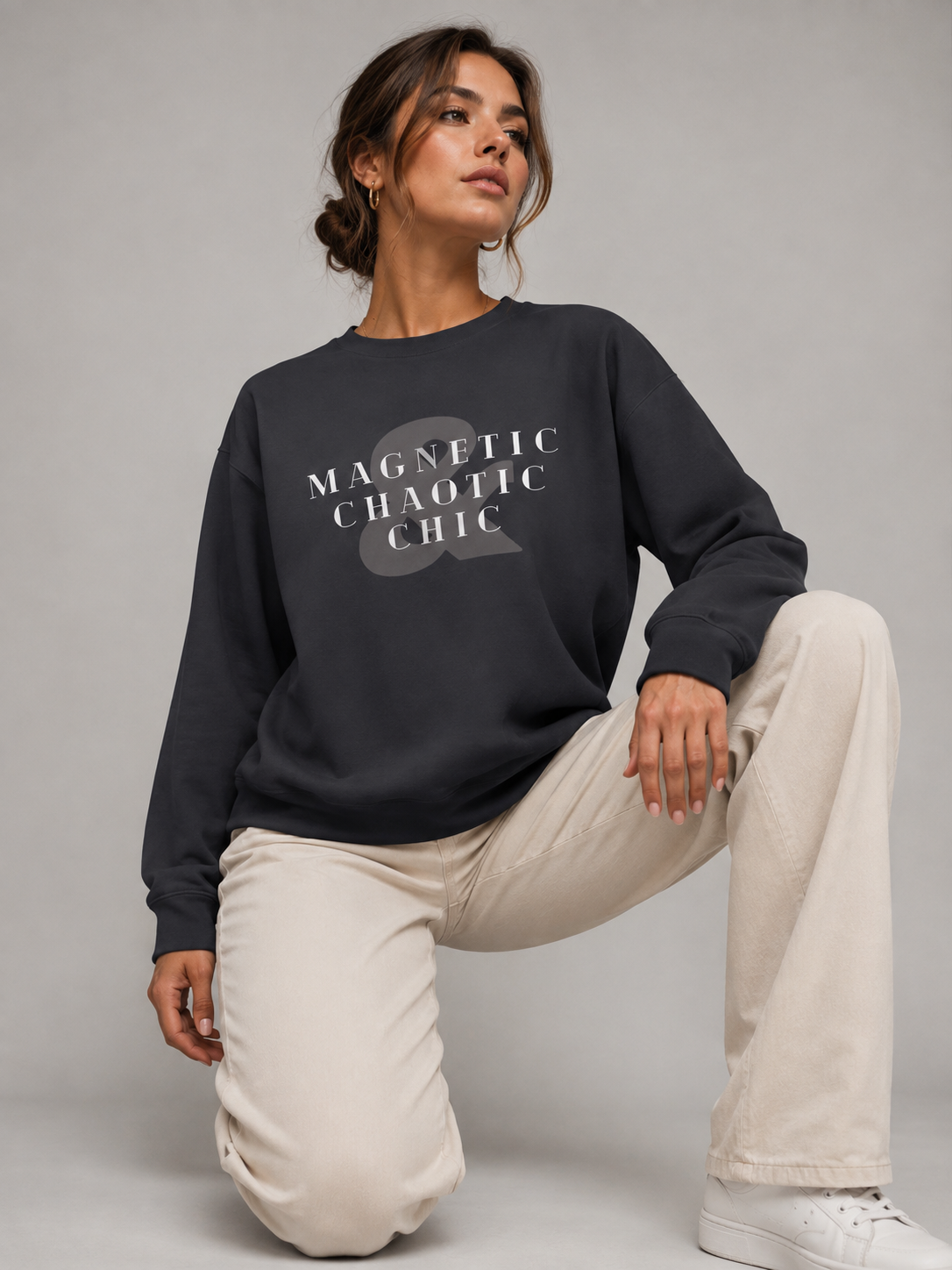 A woman wearing a dark charcoal Magnetic Chaotic Chic crewneck sweatshirt with bold white graphic typography, paired with cream trousers while kneeling on a light grey studio floor.