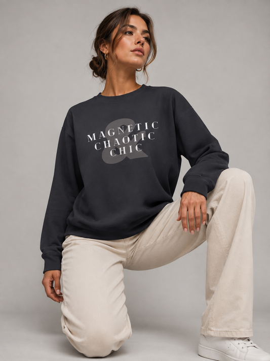 A woman wearing a dark charcoal Magnetic Chaotic Chic crewneck sweatshirt with bold white graphic typography, paired with cream trousers while kneeling on a light grey studio floor.