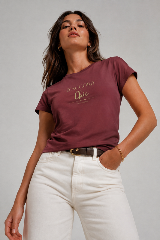 Woman wearing a burgundy D’ACCORD Chic graphic T-shirt with cream high-waisted jeans in a light gray studio, photographed from a low angle.
