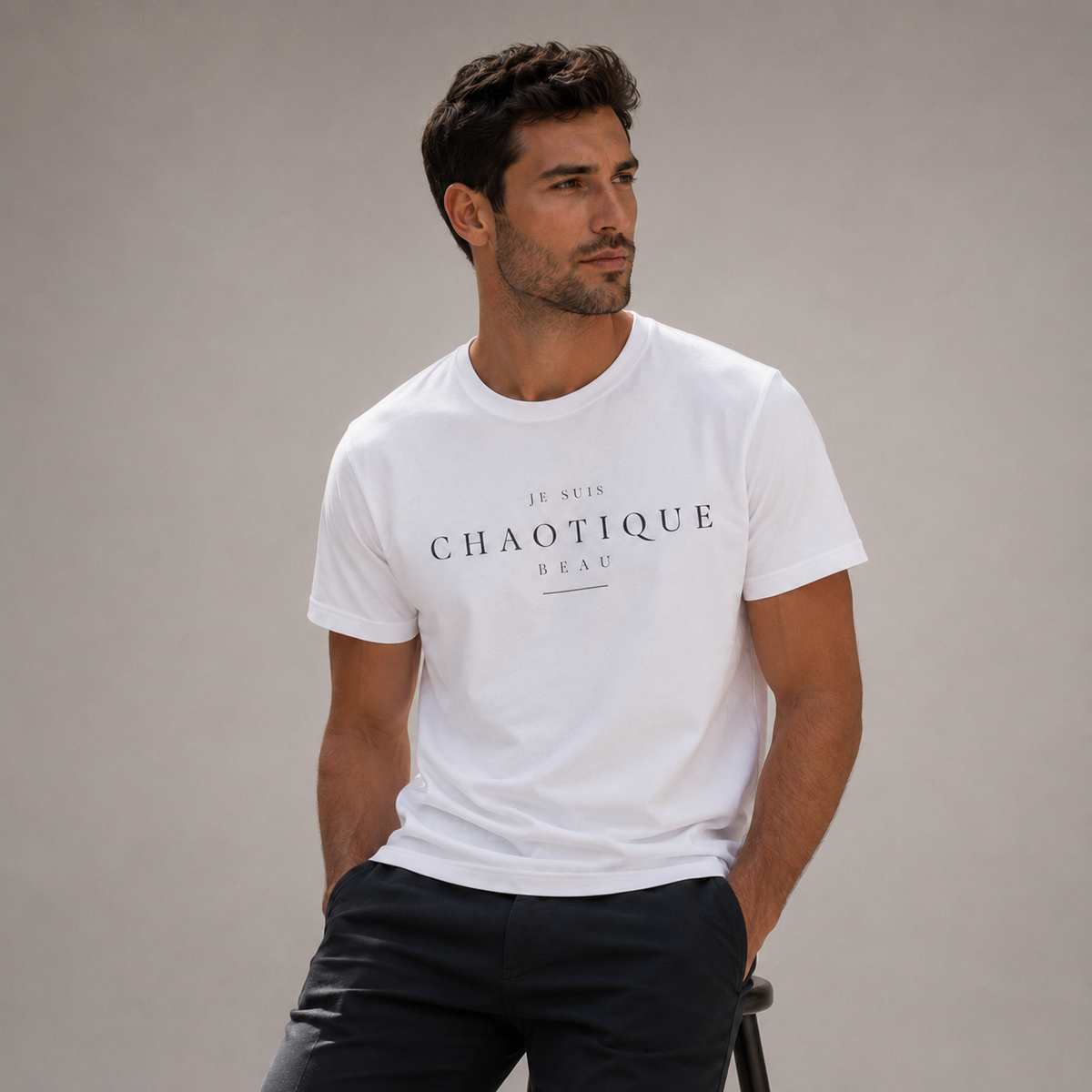 Man wearing a white ‘Je Suis Chaotique Beau’ crew-neck T-shirt with dark tailored trousers in a minimalist editorial studio setting.