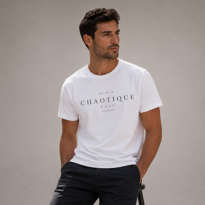 Man wearing a white ‘Je Suis Chaotique Beau’ crew-neck T-shirt with dark tailored trousers in a minimalist editorial studio setting.