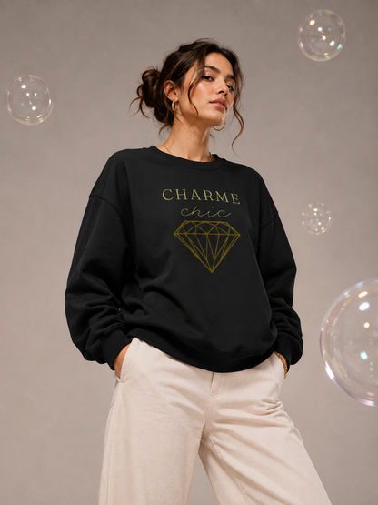 A woman wearing a black Charme Chic graphic sweatshirt with gold typography and a diamond design, paired with cream trousers in a neutral studio setting with floating iridescent bubbles.